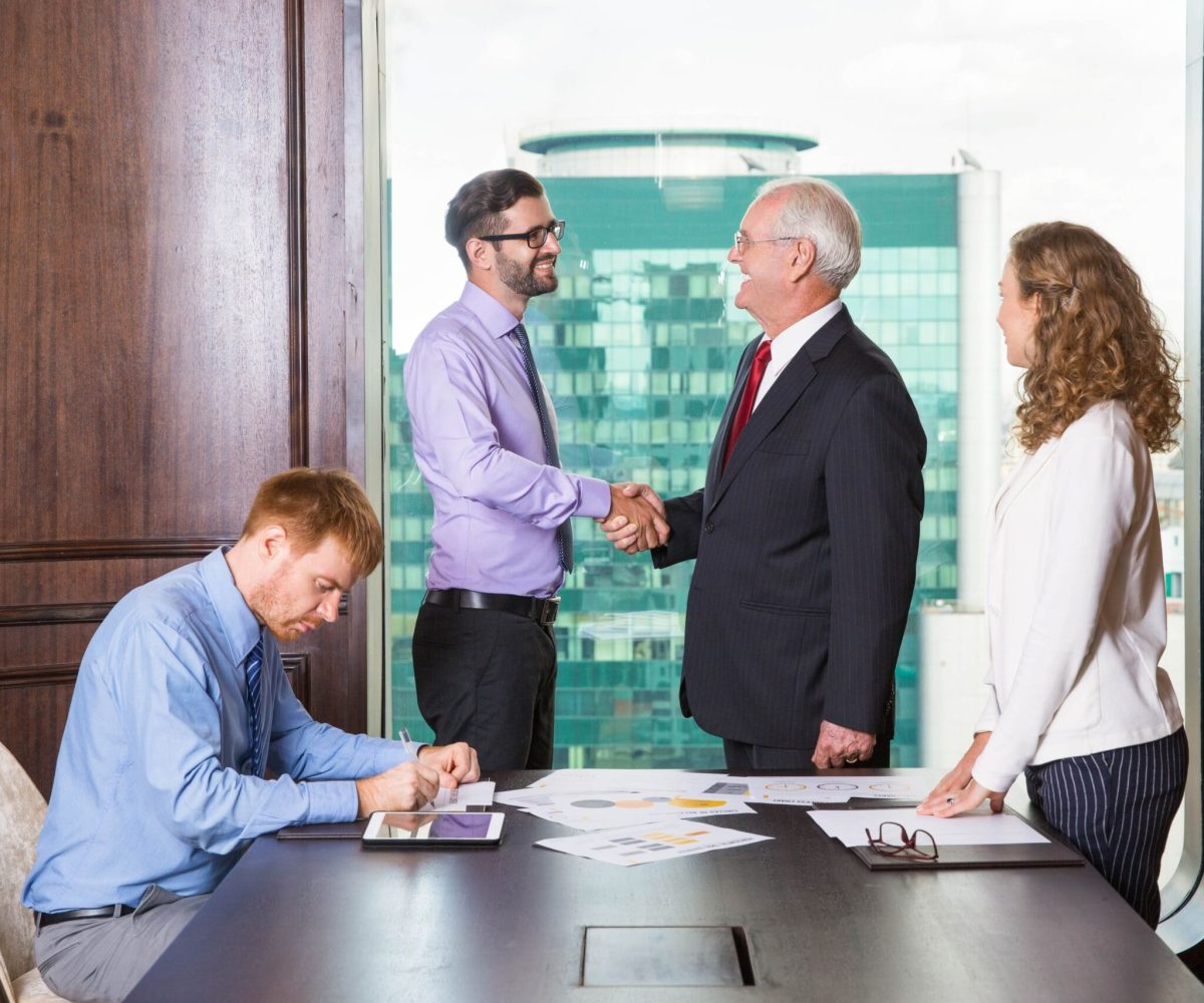 Smiling senior and young businessmen standing at head of table and shaking hands. Businesswoman is standing at table and looking at them. Another businessman is sitting at table and making notes.